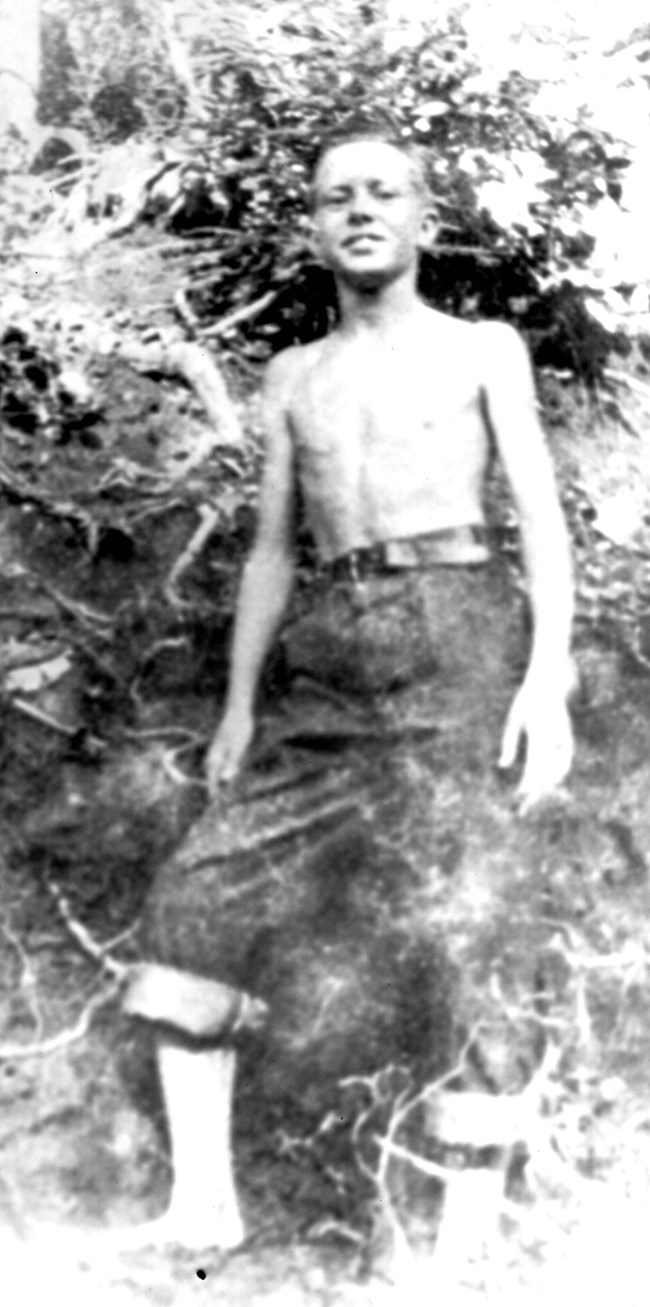 a young, shirtless Jimmy Carter stands barefooted with his pants legs rolled up. The photo was taken at FFA camp.