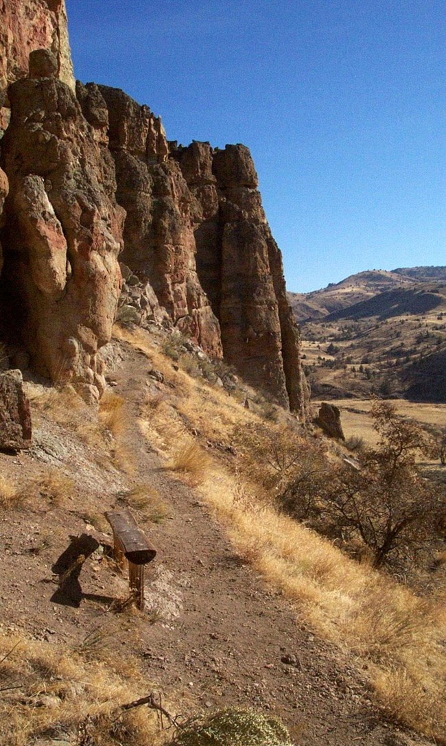 Photograph of Clarno Arch Trail featuring a clear blue sky and a gravelly trail winding along a tall, brown/red rock outcrop. Photograph of Clarno Arch Trail featuring a clear blue sky and a gravelly trail winding along a tall, brown/red rock outcrop.