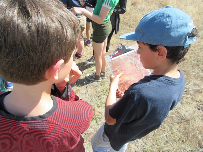 Two boys stand with the backs to the camera with clipboards in a sandy field.