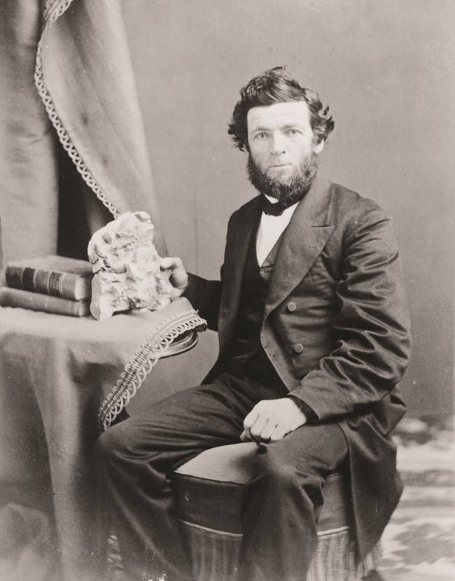 A black and white photo of a man with a beard wearing an old-fashioned suit sits at a table and holds a rock.
