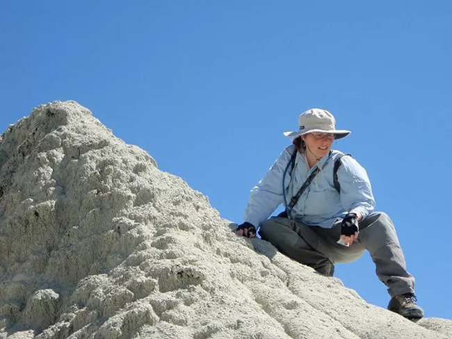 Jennifer Cavin A woman in long sleeves, pants, hiking boots, and wide brimmed hat crouches on a rock.
