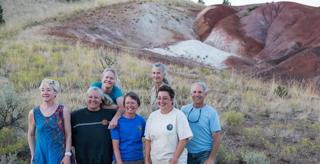 Seven smiling adult volunteers stand in front of a red hill at the Painted Hills Unit
