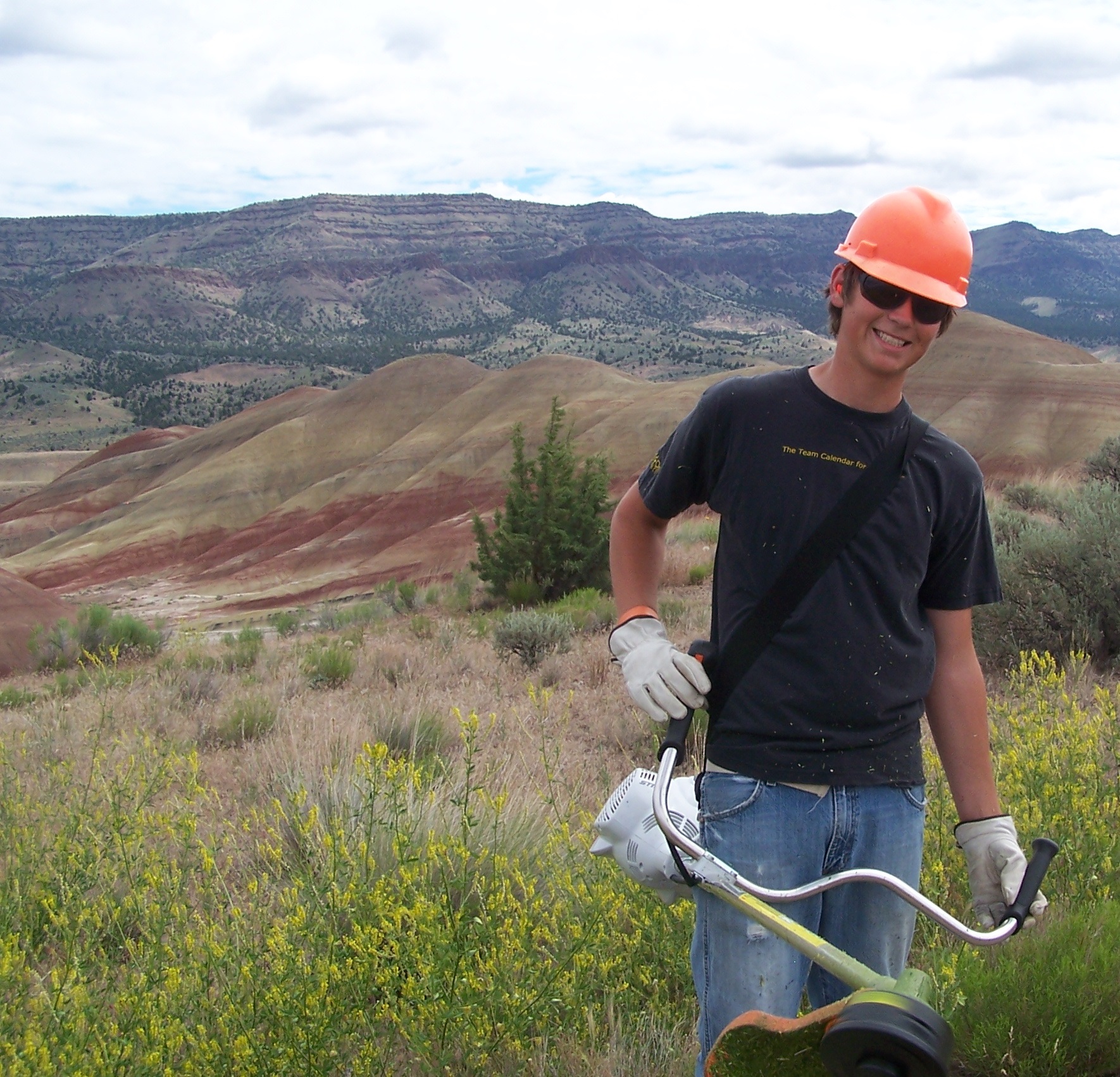 A teenager in an orage hard hat stands in the right foreground holding a weedeater. The striped hills of the Painted Hills can be seen in the right and middle of the photgraph in the background.