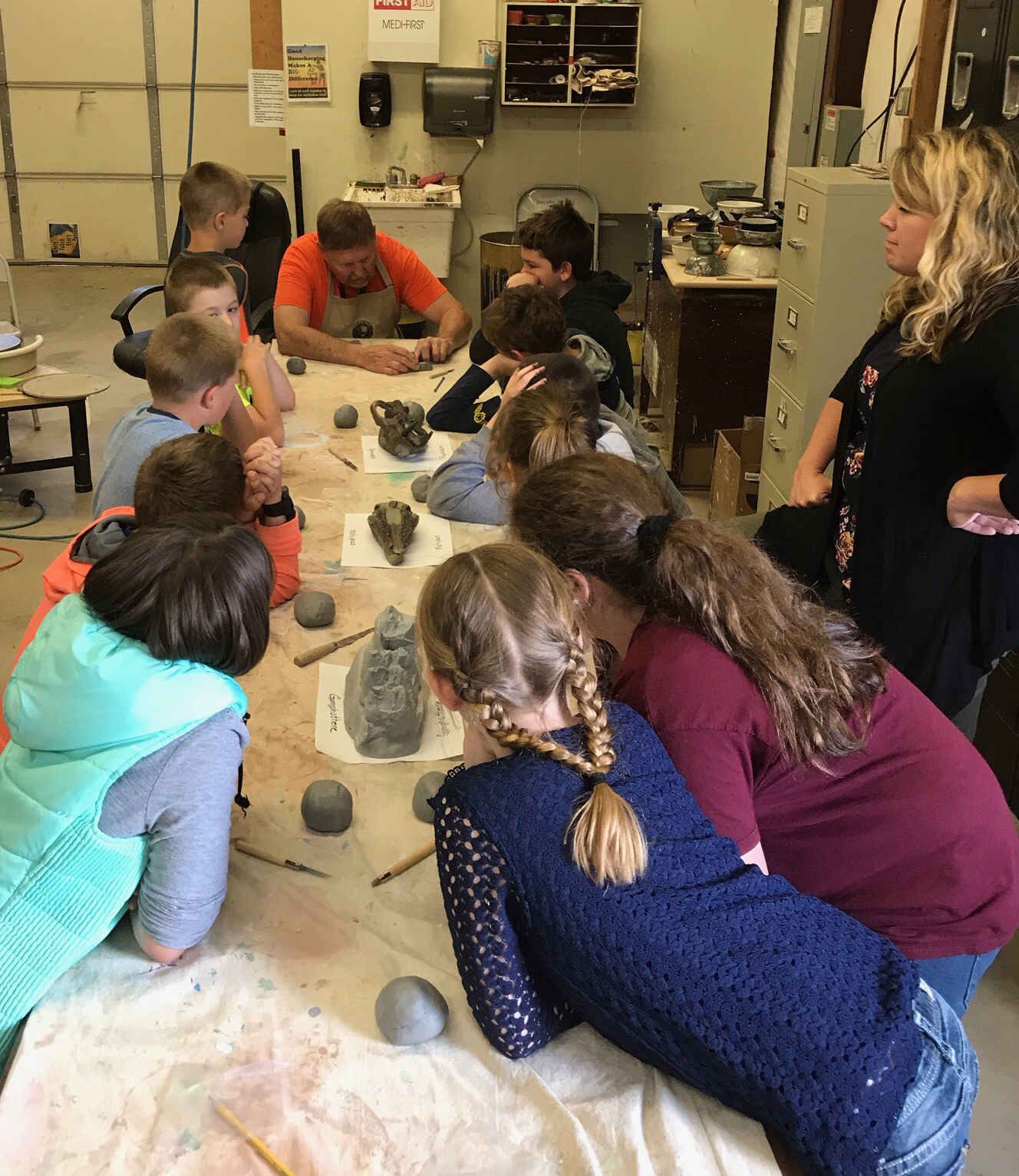 Artist-in-Residence Robin Jackson teaches pottery techniques at Dayville school. Children lead over a long table looking towards a man working on clay.