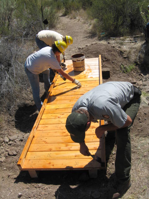 Youth Conservation Corp and Lead Three people are painting a wooden deck which is part of a trail. Two are wearing yellow hard hats and the third is wearing the green ballcap as part of the National Park Service uniform.