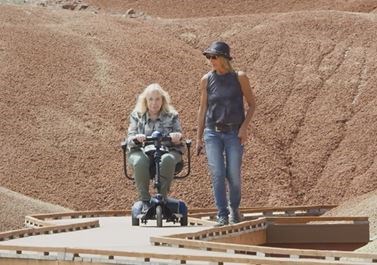 Two visitors on a boardwalk between red clay soil hills. One visitor is using an electric scooter.