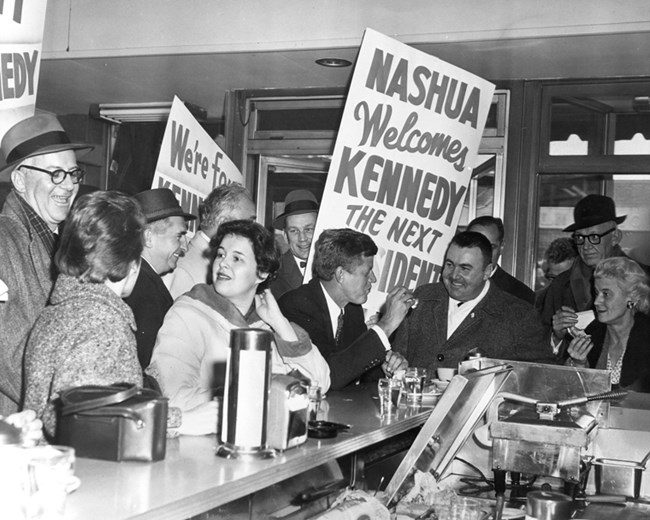 A black and white photo of JFK seated at a lunch counter with a crowd of supporters around him.