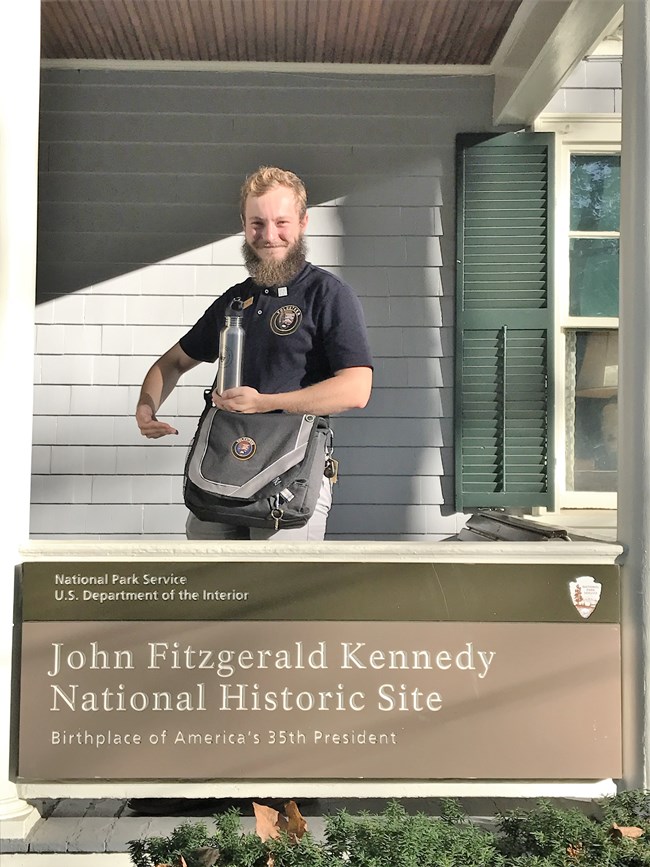 A volunteer proudly displays his volunteer uniform and traveler's bag on the porch at JFK NHS