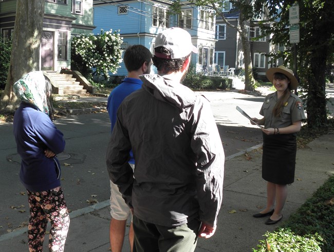 A uniformed park ranger at the right leads a group of visitors through the neighborhood.  She is holding a replica historic photograph.