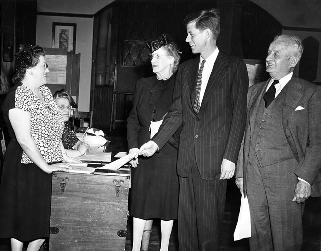 A black and white photo of JFK, flanked by his grandparents Fitzgerald, sliding his ballot into a ballot box.