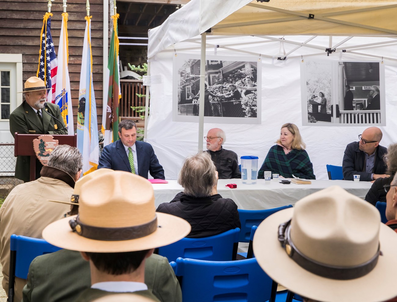 Historians sit at a table in the backyard of the John Fitzgerald Kennedy National Historic Site. Ranger Jim Roberts stands at a podium with flags behind.