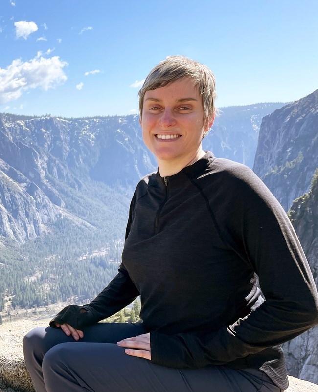 A woman in dark clothing sits and faces the camera with a mountain scene behind her