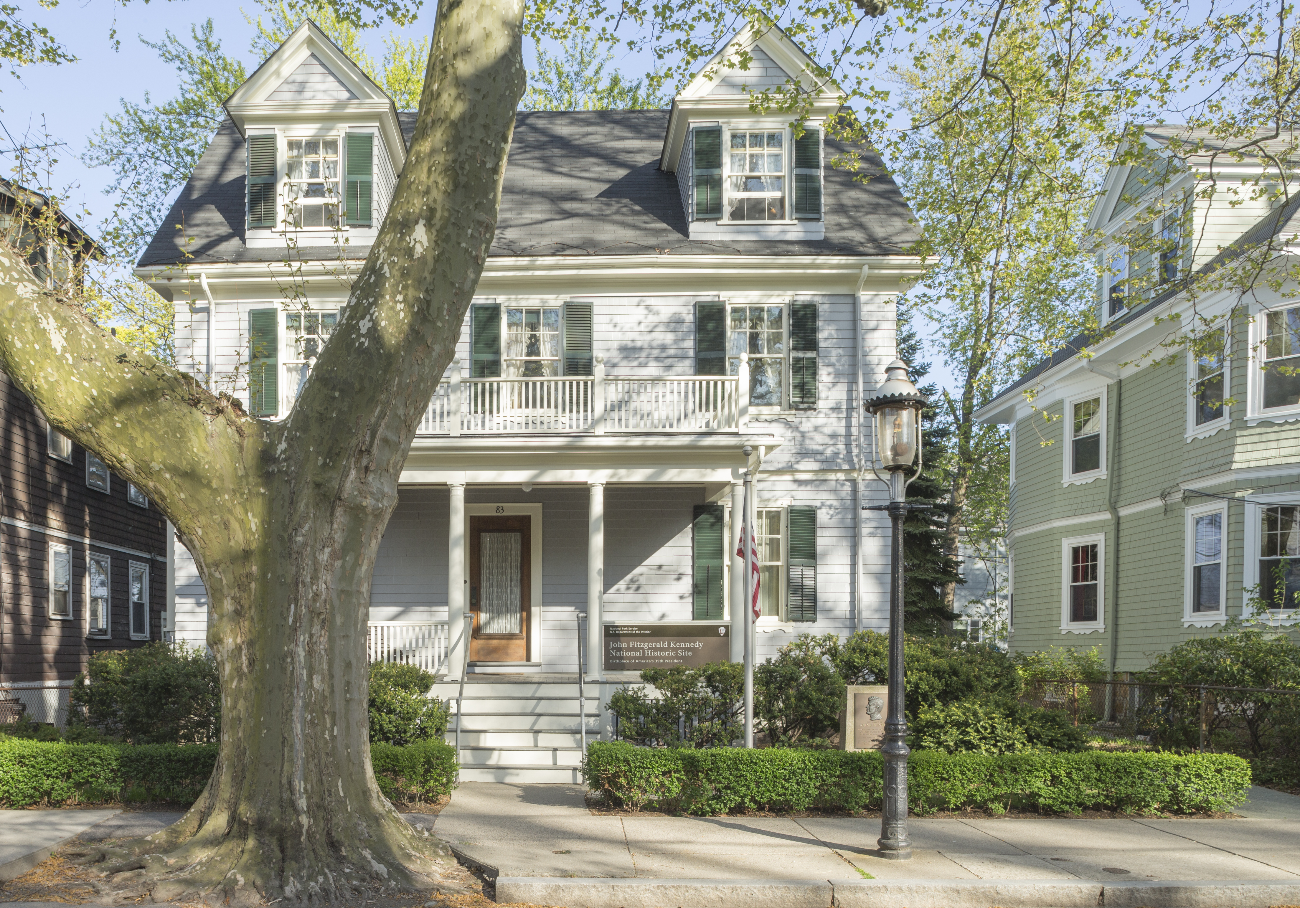 The front of a grey, two story house, partially obscured by a tree, viewed from the street.