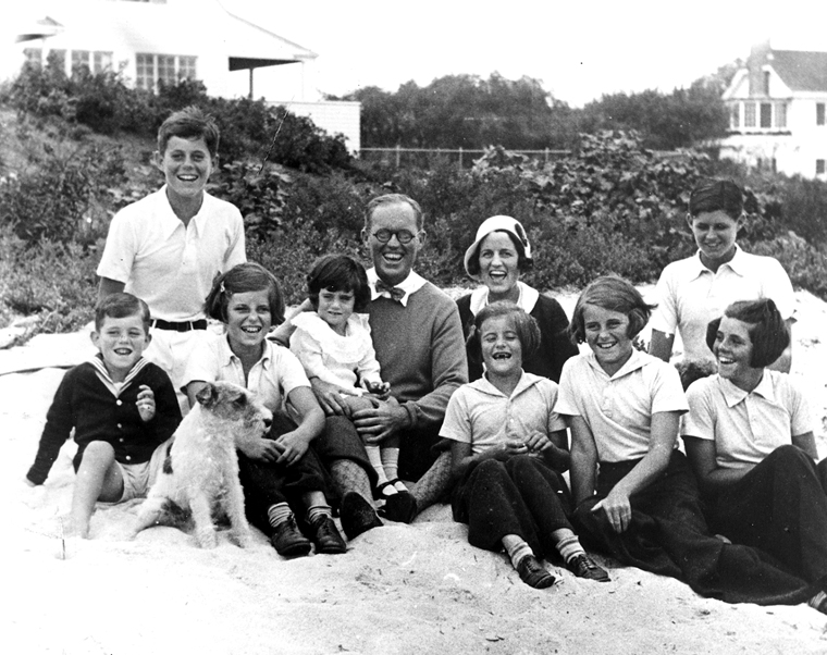 Mr. and Mrs. Kennedy pose with their eight children on a beach. They are dressed semi formally, most of them wearing matching white polo shirts.