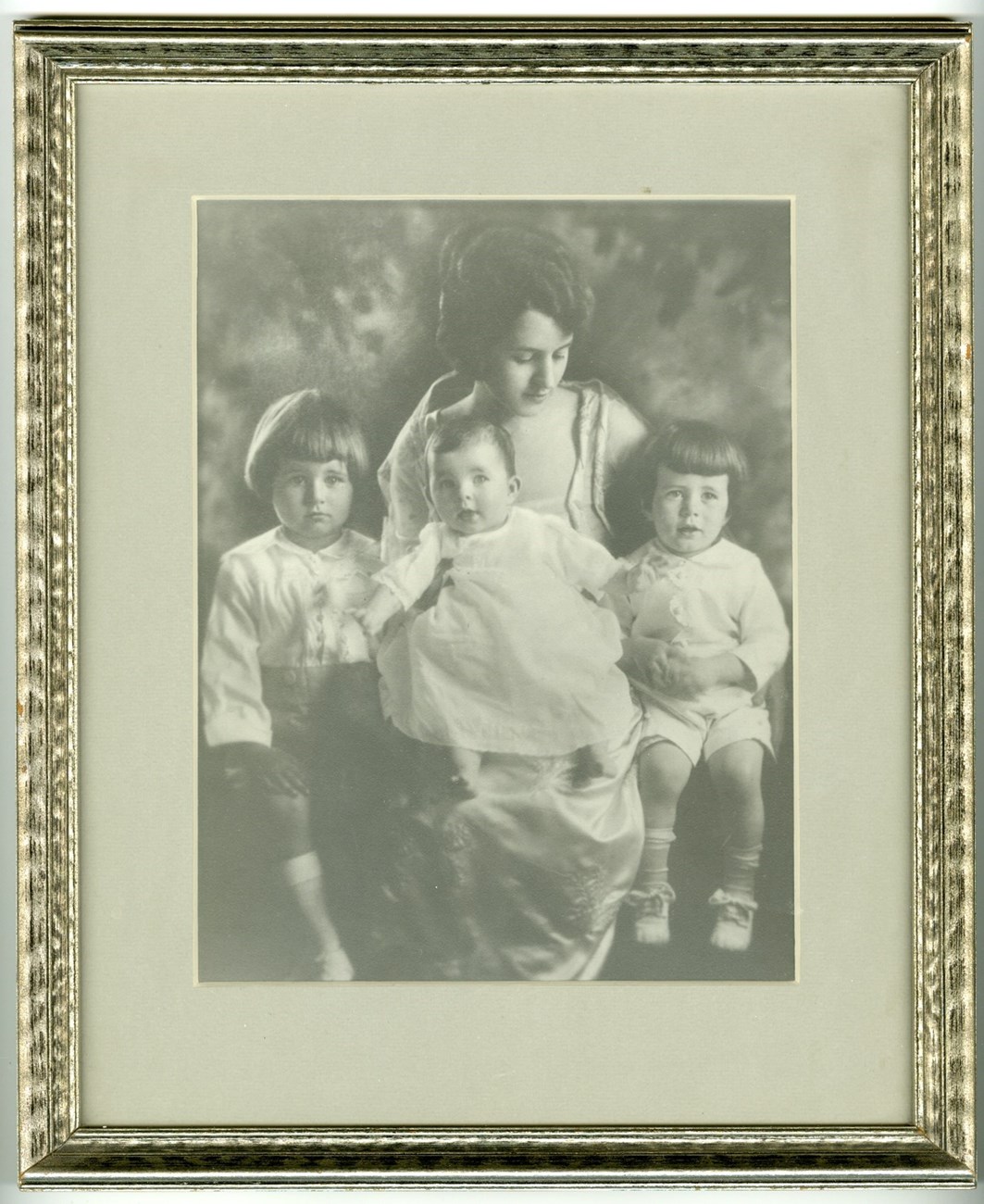 A framed black and white photograph of a young, formally dressed Rose Kennedy holding her baby on her lap and her two small children at her side, all three children also formally dressed.