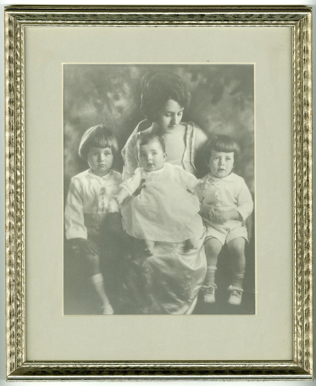 Rose Kennedy and children, ca. 1919 A framed black and white photograph of a young, formally dressed Rose Kennedy holding her baby on her lap and her two small children at her side, all three children also formally dressed.