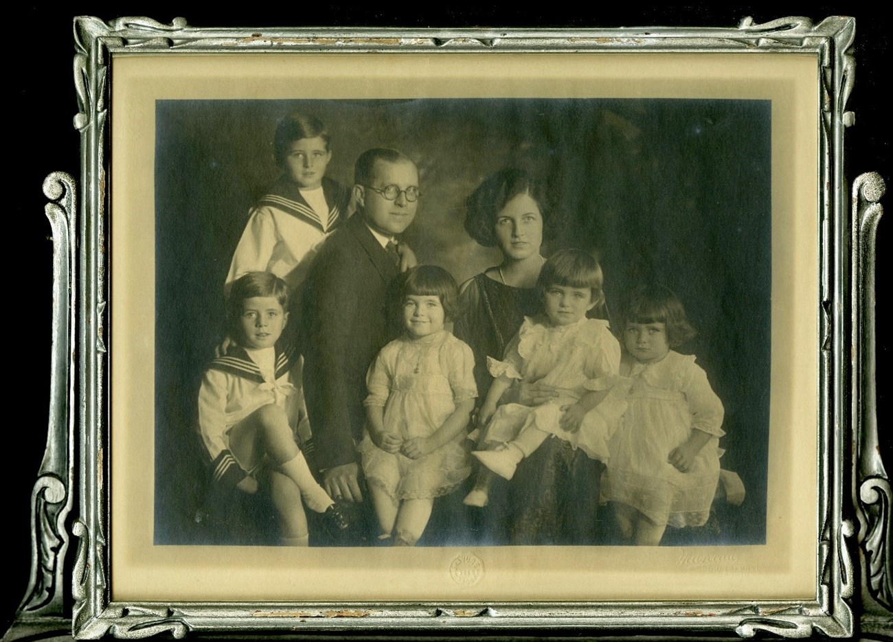 Framed photograph of formally dressed Mr. and Mrs. Kennedy and five children. From left to right, two boys in matching suits sit and stand by their father, while three daughters in matching dresses sit on their parents' laps and next to their mother.