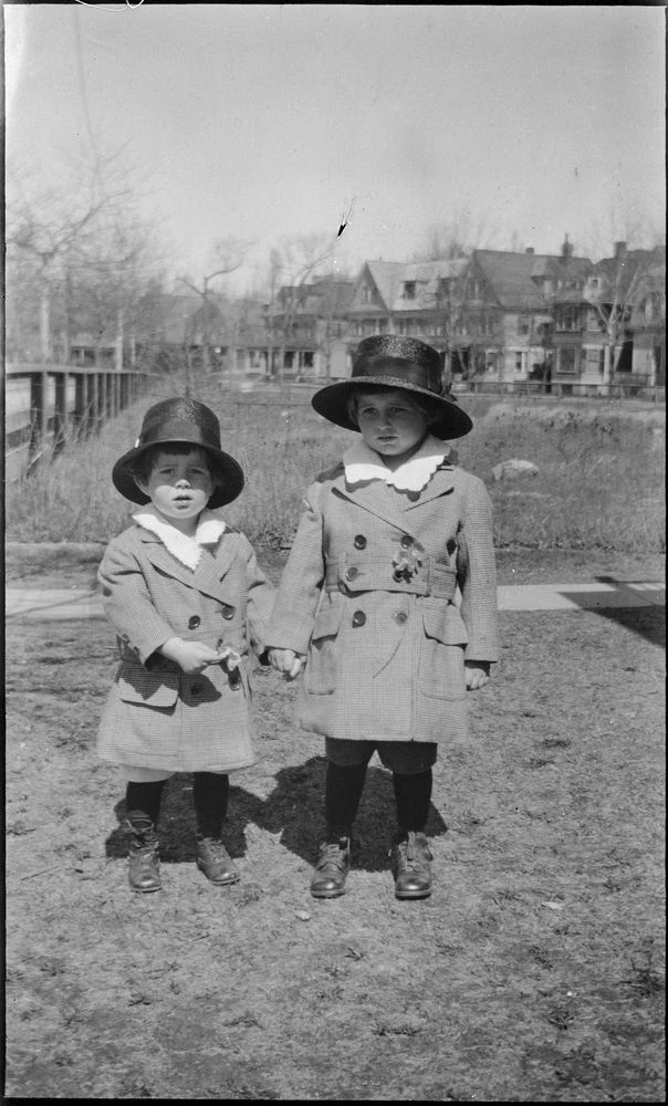 Joseph P. Kennedy Jr. and John F. Kennedy, ca. 1919 Two small boys stand outside in matching autumn clothing. The smaller boy points to his hand, being held by the bigger boy.