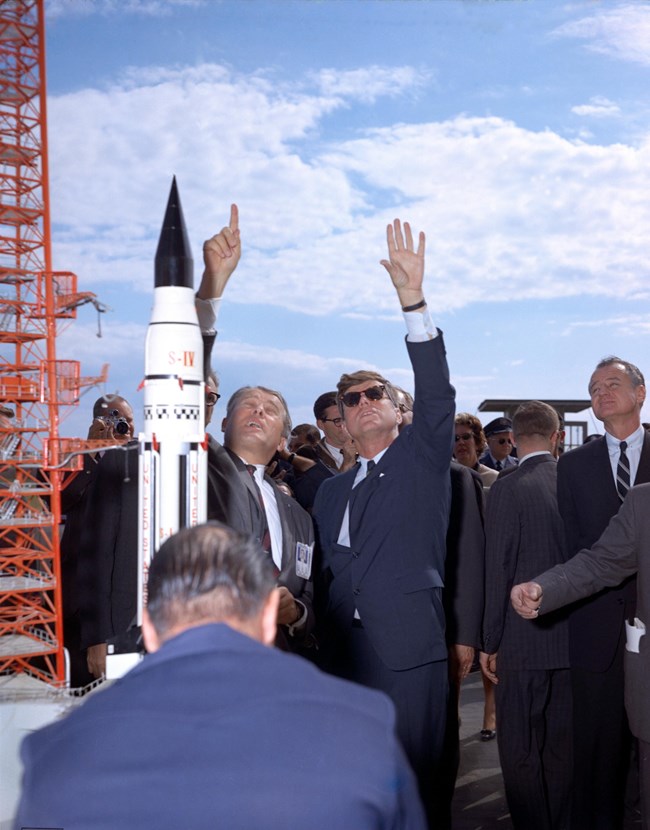 (L-R) Dr. Wernher von Braun, the NASA director of the Marshall Space Flight Center, and President Kennedy at Cape Canaveral, FL. 1963.  Each man is looking up pointing to the blue sky above.  A Saturn Rocket is on the left of the photo.