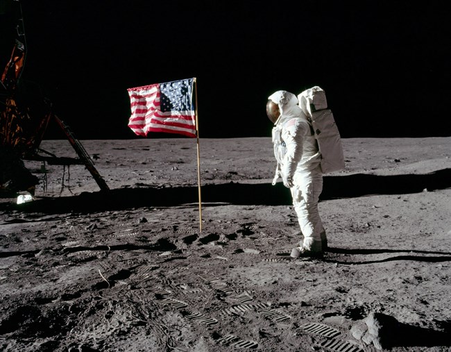 Astronaut Neil Armstrong stands on the moon.  Armstrong is on the right side of the photo.  He is wearing a white space suit with a tank on his back.  He looks at an American flag.  He is standing on the crater-filled gray landscape of the moon.