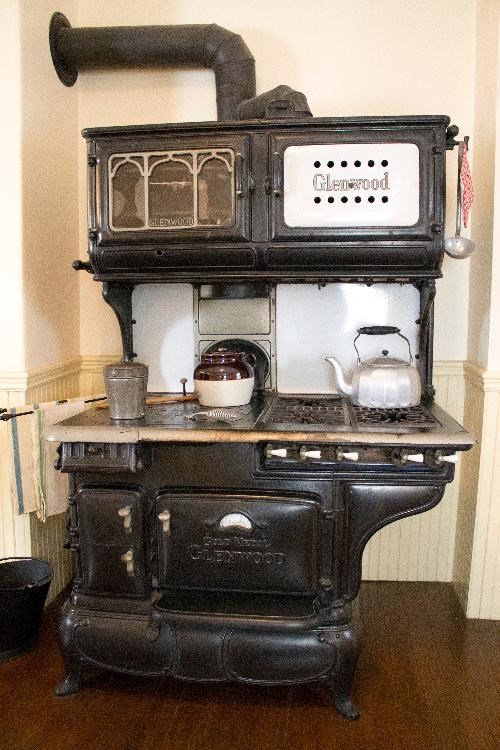 A Glenwood gas and coal burning stove.  The stove is black and has a variety of oven doors.  There are several items atop the stove, including a silver kettle and ceramic bean pot.