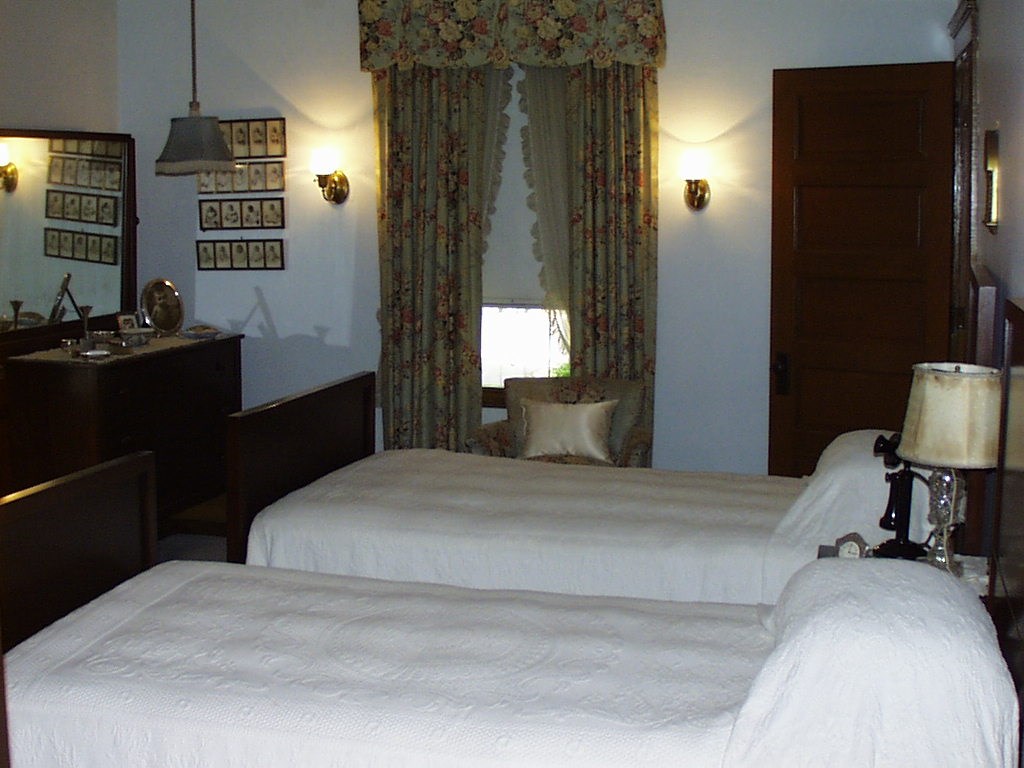Two twin beds covered with white embroidered spreads fill the room.  A window and baby photos are at the far end of the room.  A dark wooden dresser stands to the left of the beds.