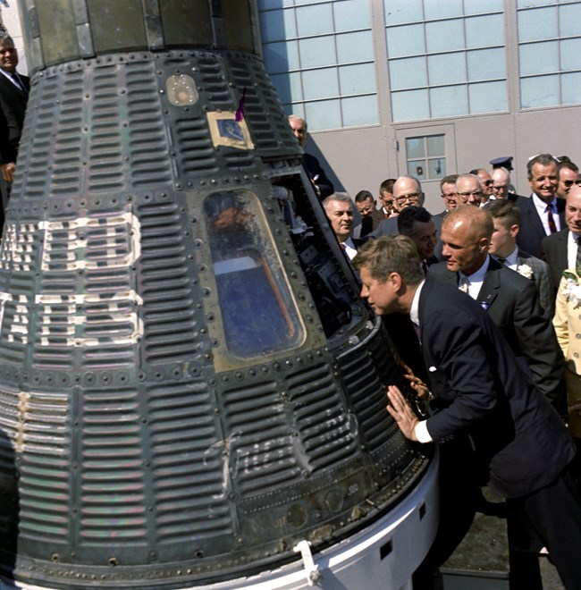 President Kennedy and astronaut John Glenn Jr. look inside space capsule Friendship 7. 1962.  Both men are wearing dark suits and staring into the capsule from the right.  A group of people are standing behind them.