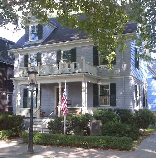 Grey house on a tree-lined street. US Flag on pole.
