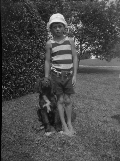 A young JFK stands on grass with his dog.  The photo is black and white.  JFK is wearing a striped shirt, shorts, and a sun hat.  The dog is a black spaniel.