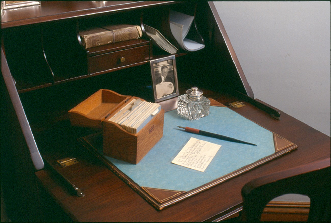 A brown wooden desk.  On the top sits a wooden card file box, a card on JFK, a fountain pen, ink well, and a photograph of Rose's father, John F. Fitzgerald, holding Joe Jr.