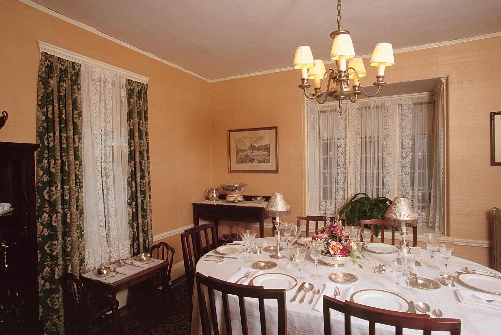 A large dinner table sits in the center of the room.  It is topped with fine dinnerware.  Above the table hangs a chandelier.  By the left window, sits a children's table.