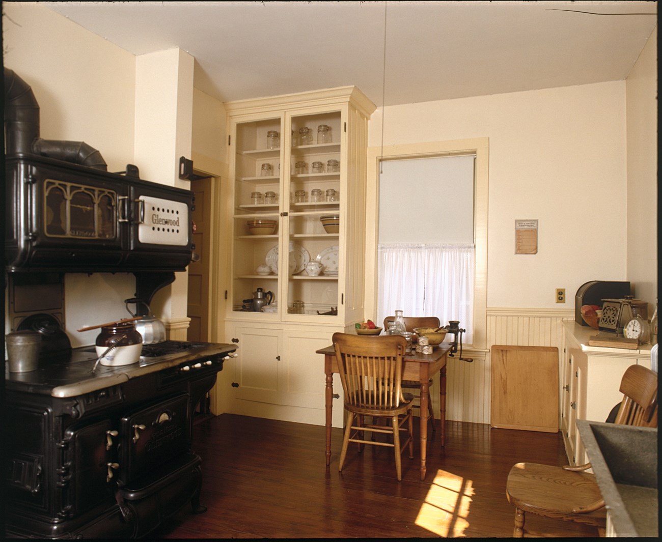 A black Glenwood coal and gas burning stove is on the left.  A white cabinet with glass doors houses cookware.  A small wooden table sits at the back of the room.  On the right is a counter with a soapstone sink.