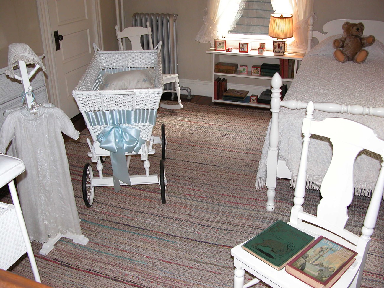 A white bassinet and christening gown at the left of the photo.  A white chair with two children's books is at the right forefront.  Behind the chair is a white twin bed with a teddy bear on top.  A white bookcase sits to the left of the bed.