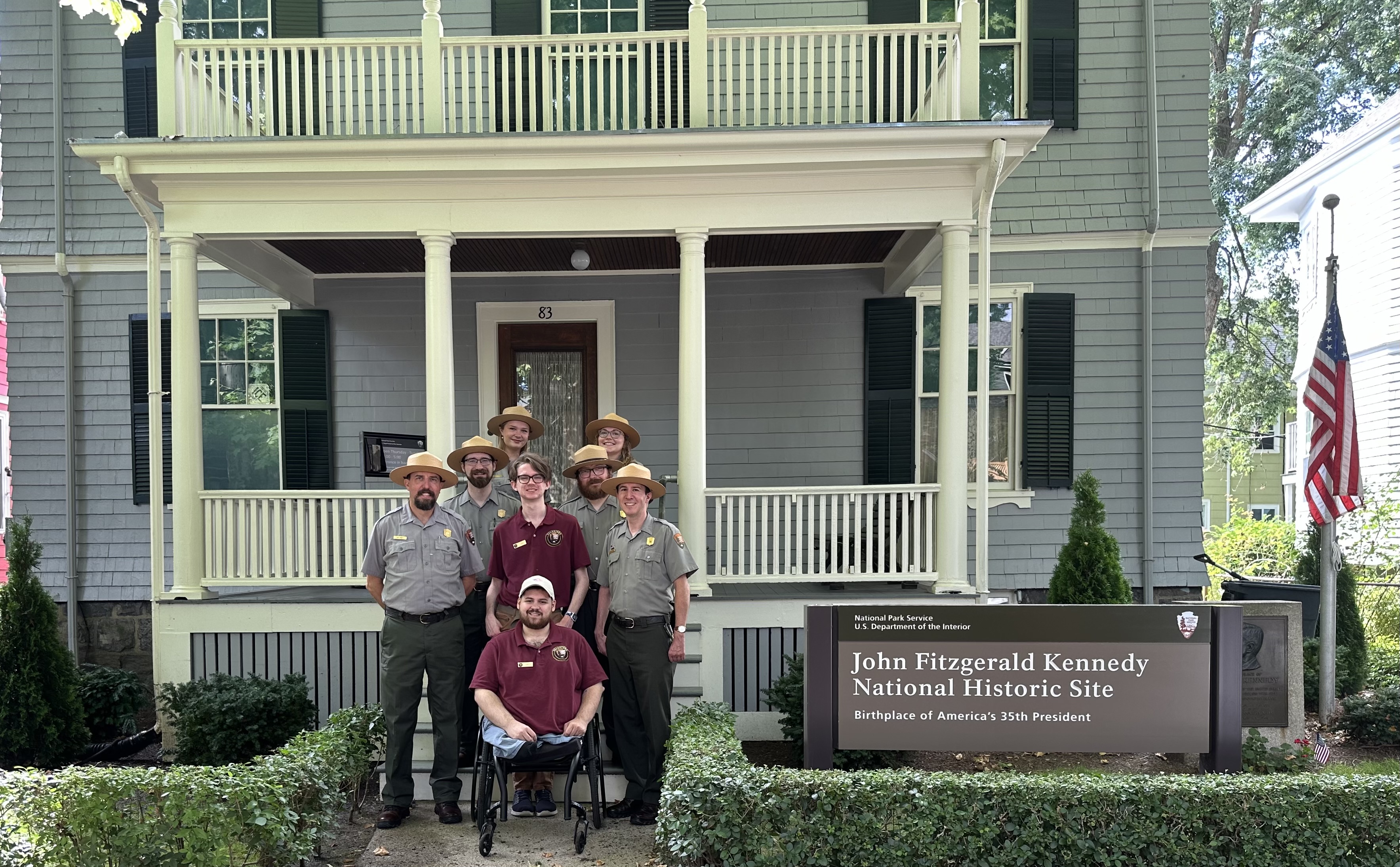 A group of people are facing the camera in front of a gray house with dark green shutters
