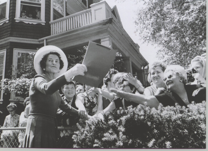 A woman in a hat and dress hands and envelope to a crowd of women.