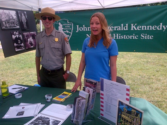 A uniformed park ranger and an intern stand behind a table with brochures.  A banner reading John Fitzgerald Kennedy National Historic Site appears in the background with photos of the Kennedy family.