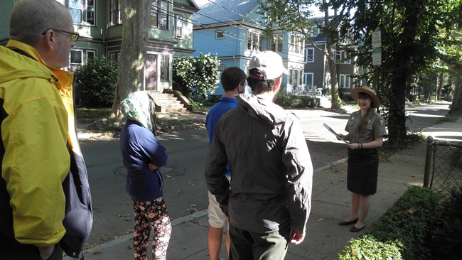 A park ranger guides visitors along a sidewalk.  She is holding several historic photos