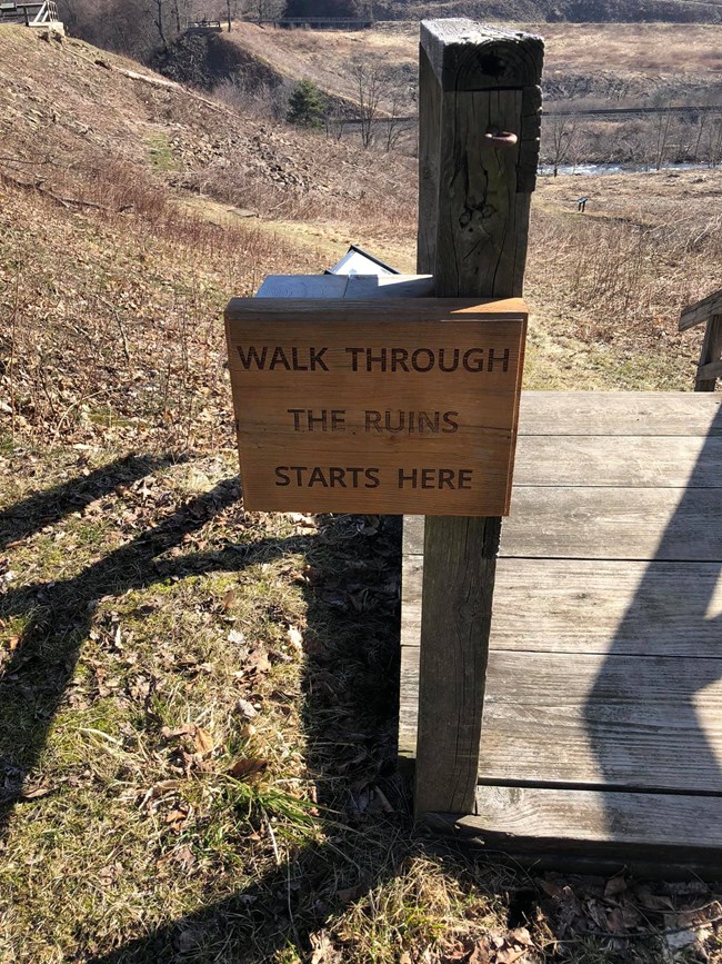 A sign that says, a walk through the ruins starts here with the remains of the South Fork Dam in the background.