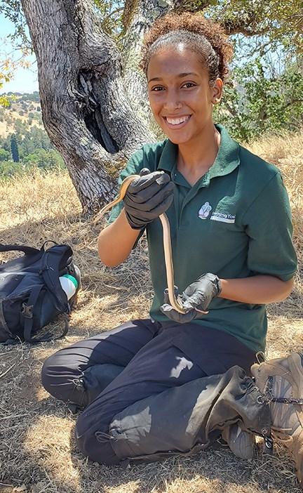 A young female of color, in a polo shirt is holding a small snake, outdoors. She is smiling at the camera. Trees and grass surround her.