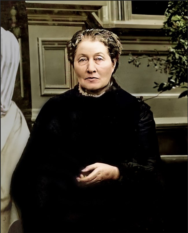A portrait of Louisa 'Louie' Strentzel Muir, John Muir's wife, seated in front of a building with a composed expression. She is dressed in dark clothing.