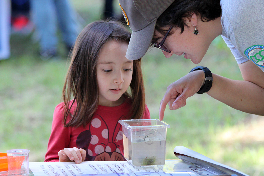 Little girl looks in a plant specimen container as a female volunteer describes its content.