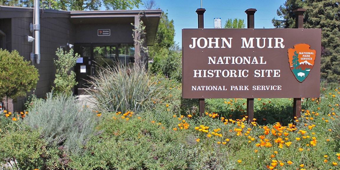 A wooden park sign, with park name, in front of the John Muir Visitor Center.