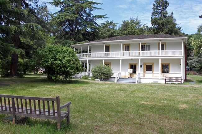 The two story adobe home sits amongst the grass and trees. A bench can be seen in the foreground.