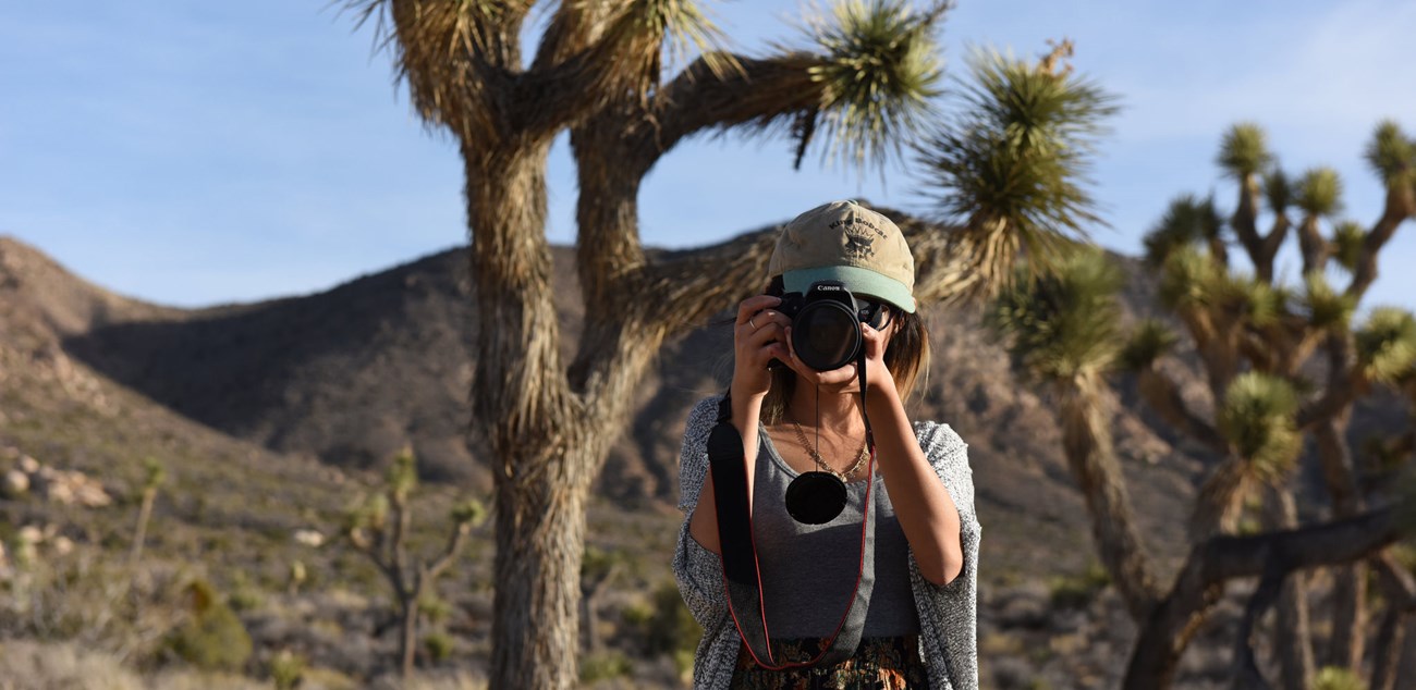 a woman wearing a hat holds a large camera to her eye pointed directly at you with mountain slopes and spindly Joshua trees behind her