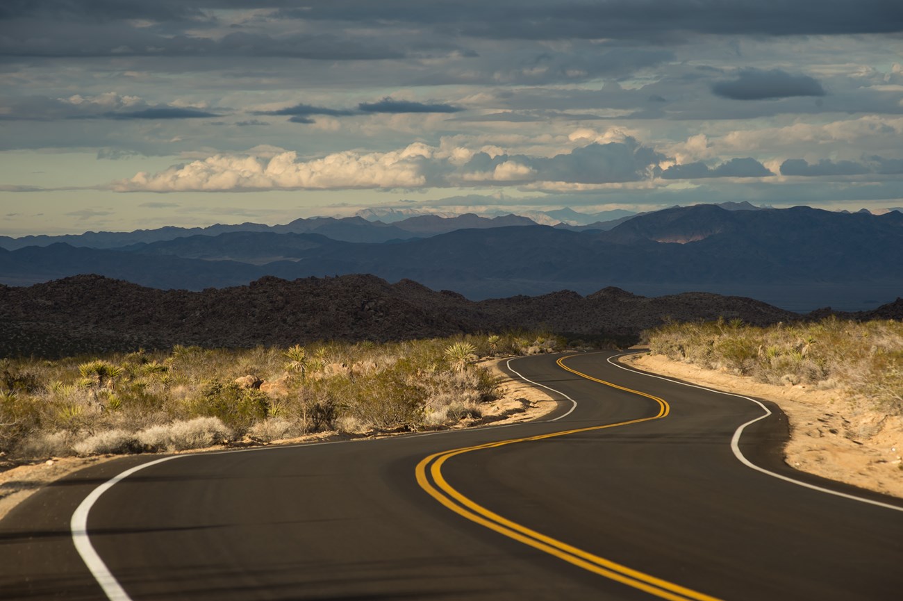 A paved road winds through a desert landscape under a cloudy sky.