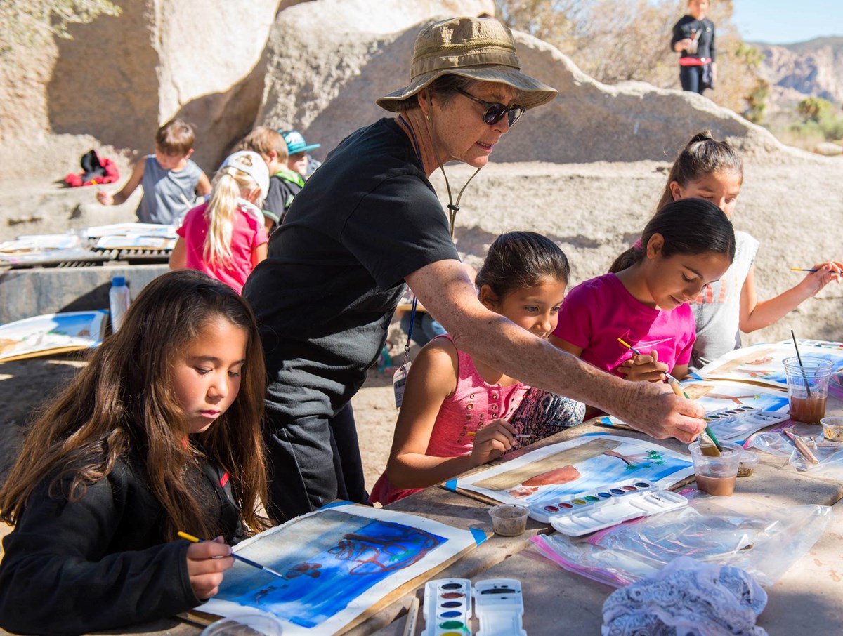 Color photograph of four children and adult artist in the foreground painting nature scenes with watercolors. In the background other children can be seen painting and playing on large boulders. Color photograph of four children and adult artist in the foreground painting nature scenes with watercolors. In the background other children can be seen painting and playing on large boulders.