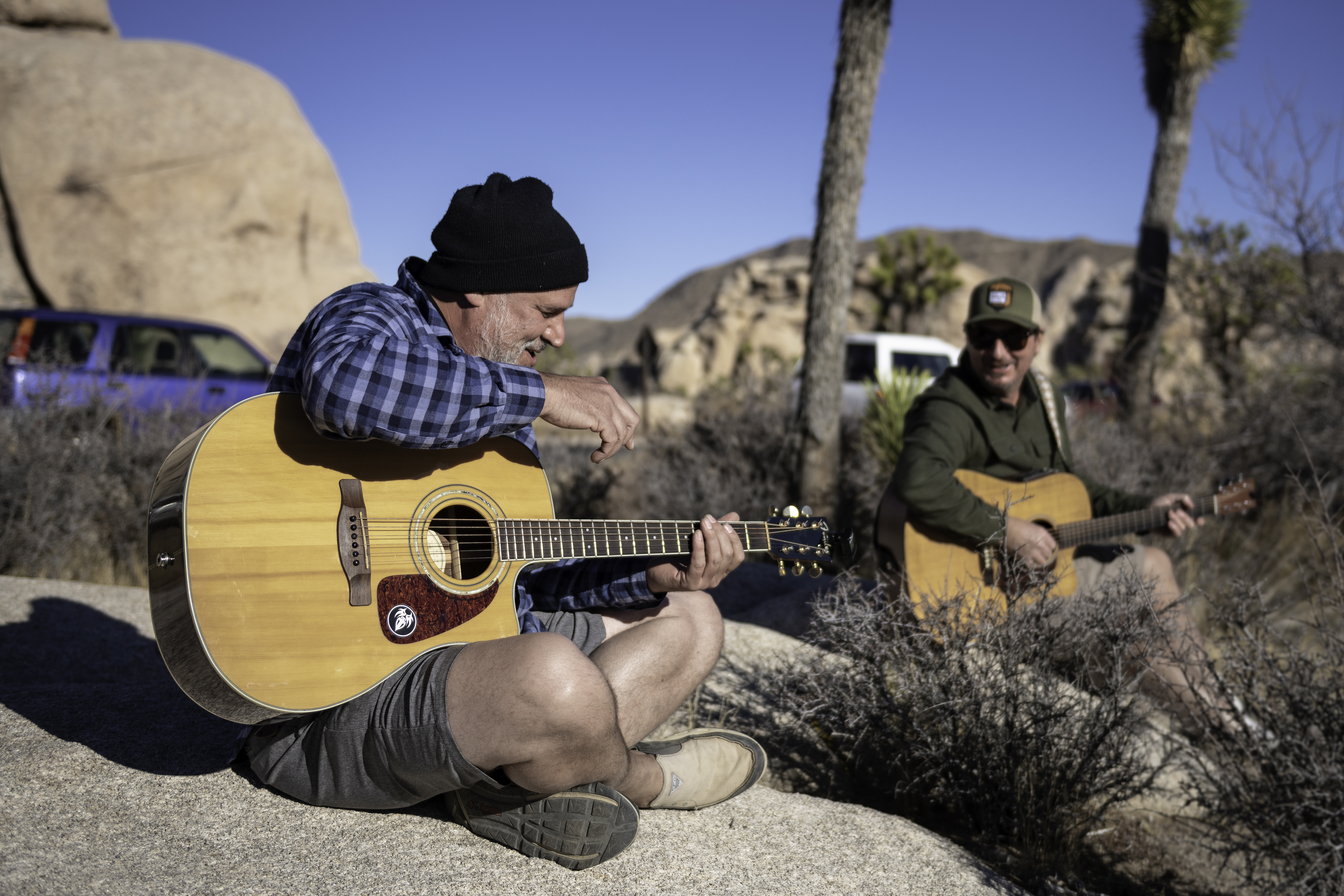 Two men sit on a rock and strum guitar.