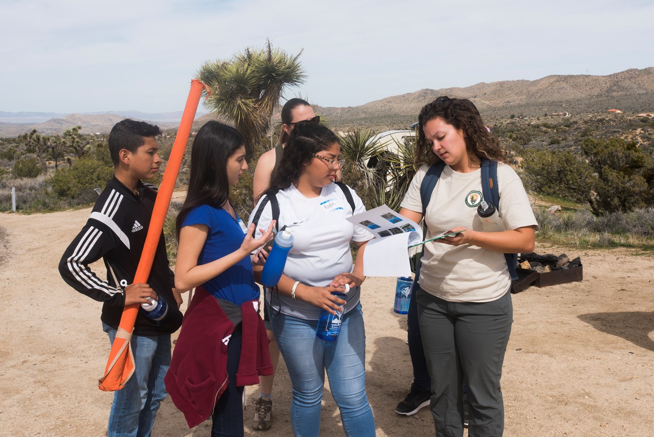 Adult woman in t shirt with park service logo shows three high school students papers on a clipboard.