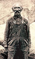 Sepia-toned photo of Johnny Lang standing in a pair of overalls and a button-down shirt. Johnny is bald with an epic mustache.