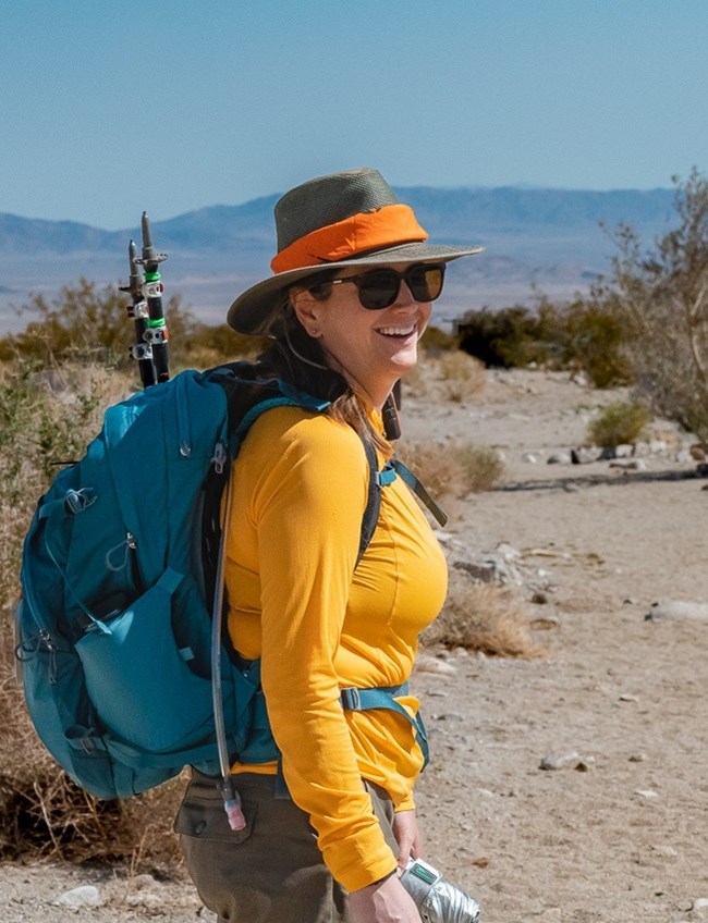 A person with a sunshirt, hat, sunglasses, and backpack smiles at the camera